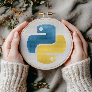 May include: A cross-stitch embroidery hoop featuring the Python programming language logo in blue and yellow. The design is set against a white fabric background, framed by a wooden hoop. The hoop is held by a person wearing a cream-colored sweater.