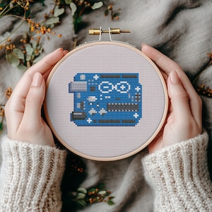 May include: A cross-stitch embroidery of an Arduino circuit board in a wooden hoop. The design features a blue circuit board with white and gray details. The hoop is held by hands, with a neutral background.