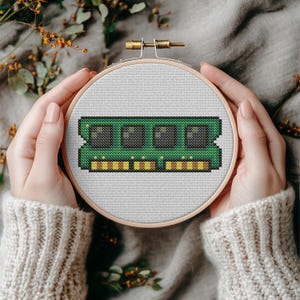 May include: A circular wooden embroidery hoop containing a cross-stitch pattern of a green computer RAM memory module. The design features four black square chips and a row of yellow and brown rectangular contact pins on a white fabric background.