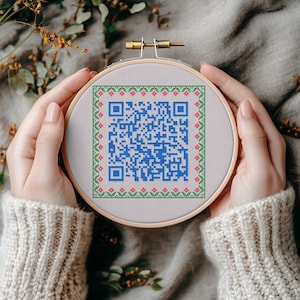 May include: A circular wooden embroidery hoop containing a light grey fabric with a cross-stitched blue QR code in the centre. The QR code is framed by a decorative border featuring small green geometric patterns and tiny pink floral accents.