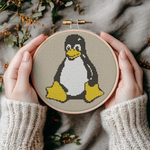 Op de afbeelding: Een borduurring met kruissteek met een pinguïnontwerp. De pinguïn is zwart, wit en geel, op een beige stof. De houten ring wordt door handen vastgehouden, met een neutraal gekleurde trui op de achtergrond. Het knutselproject is handgemaakt.