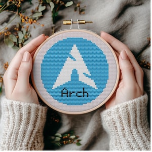 May include: A cross-stitch embroidery hoop featuring a blue circle with a white graphic and the word "Arch" in black. The embroidery is held in a wooden hoop. The background is a soft gray fabric.
