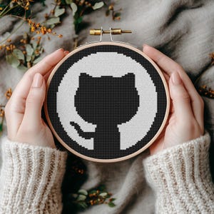 May include: A black and white cross-stitch design of a cat silhouette within a white circle, set in a wooden embroidery hoop. The design is held against a neutral-toned background with foliage.