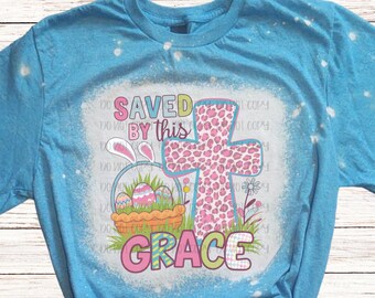 May include: A light blue t-shirt with a graphic design featuring a pink cross with a leopard print pattern, an Easter basket with eggs, and the words "SAVED BY THIS GRACE". The shirt has a bleached effect.