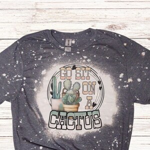 May include: A dark gray t-shirt with a bleached design featuring a cactus illustration and the text "GO SIT ON A CACTUS". The design includes a circle with cacti in pots and decorative elements. The shirt has a crew neck.