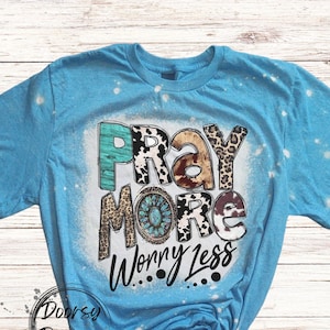May include: A light blue t-shirt with the words "PRAY MORE WORRY LESS" in a mix of fonts and patterns. The word "PRAY" is in turquoise and brown, while "MORE" features a turquoise and brown design. The shirt has a bleached effect.