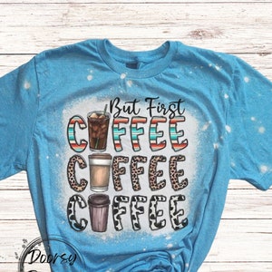 May include: A light blue t-shirt with a bleached design featuring the text "But First Coffee" and three coffee cups with different designs. The coffee cups are surrounded by a leopard print pattern.