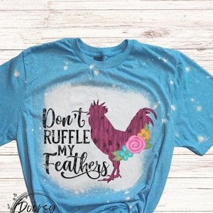 May include: A light blue t-shirt with a distressed design. The shirt features a graphic of a purple rooster with floral accents and the text "Don't Ruffle My Feathers". The shirt has a crew neck and short sleeves.