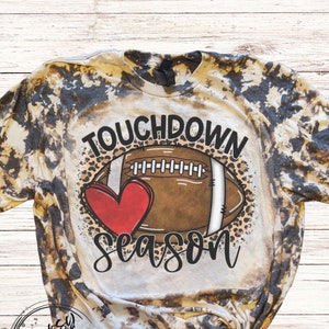 May include: A bleached t-shirt with a football-themed graphic. The design features a football, a red heart, and the words "Touchdown Season" in a script font. The shirt has a brown and tan tie-dye pattern.