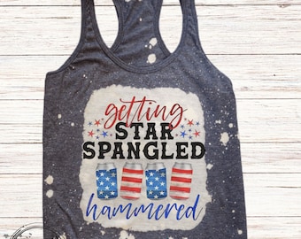 May include: A navy blue racerback tank top with a bleached white design. The text "getting star spangled hammered" is printed on the front, along with images of beer cans in red, white, and blue. Fireworks are in the foreground.