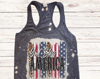 May include: A blue tank top with a distressed American flag design and the text "God Bless America" in white. The flag is layered with a leopard print pattern. The background is a white wood surface.