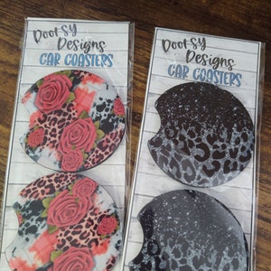 May include: Two sets of car coasters featuring a leopard print design. One set has a black and white leopard print with a white splatter effect. The other set has a pink, red, and white floral design with a leopard print background. Both sets have the text "Boot-sy Designs Car Coasters" on the packaging.