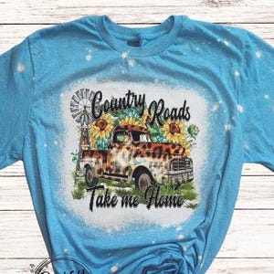 May include: A light blue t-shirt with a graphic featuring a vintage truck, sunflowers, and the text "Country Roads Take me Home." The shirt has a bleached, distressed look, with white spots scattered across the fabric. The design includes a windmill.