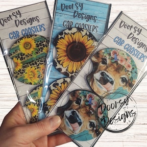 May include: Three sets of car coasters featuring different designs. The first set features a sunflower design with a black and white cow print background. The second set features a cow with a floral headband and a blue background. The third set features a sunflower design with a black and white cow print background. All sets have the text "Doot Sy Designs CAR COASTERS" printed on them.