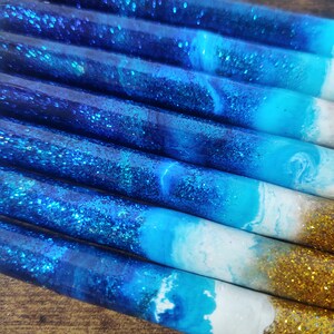 May include: Close-up of several blue and white resin pens with gold glitter accents. The pens have a gradient of blue and white colors, resembling a beach scene.