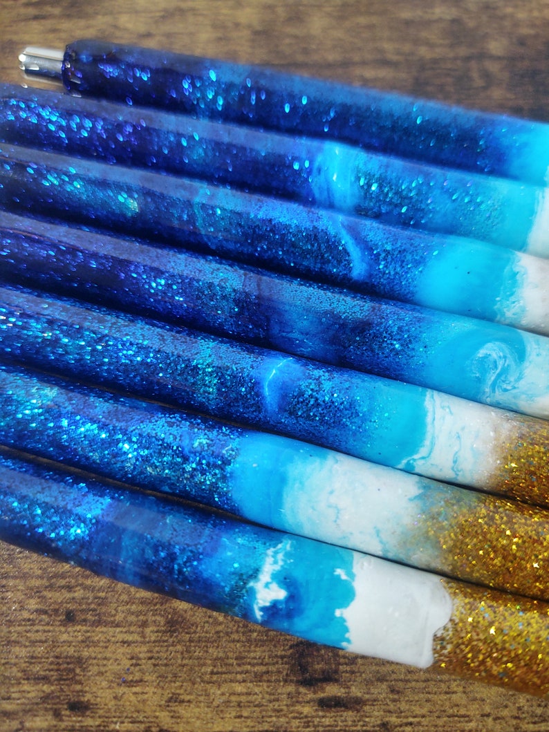 May include: Close-up of several blue and white resin pens with gold glitter accents. The pens have a gradient of blue and white colors, resembling a beach scene.