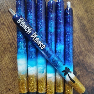 May include: Set of seven blue and gold glitter pens with a beach theme. The pens have a silver top and a black tip. One pen has the text "Beach Please" written on it.