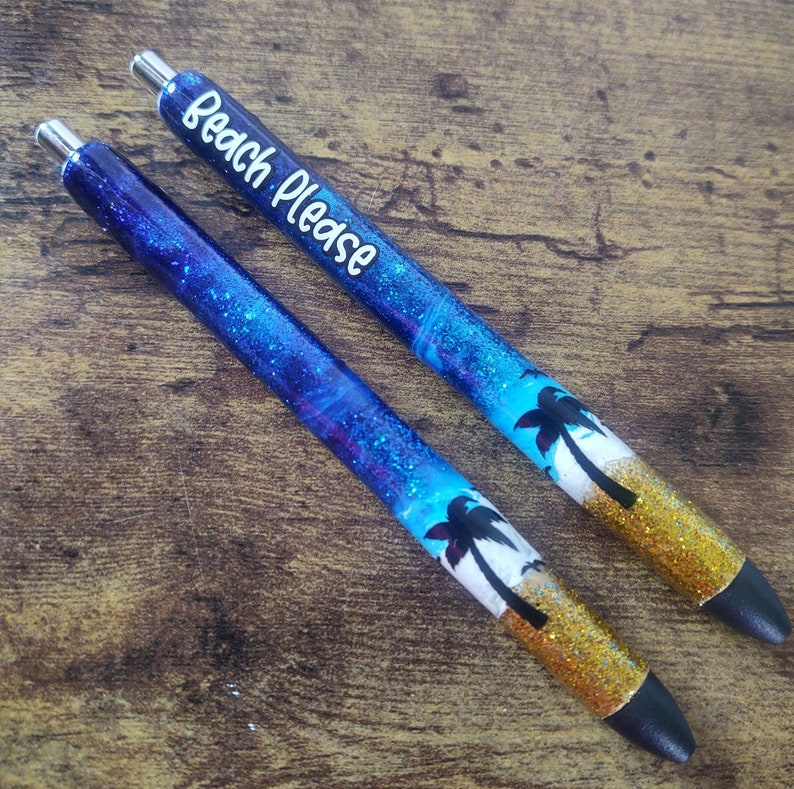 May include: Two blue and gold glitter pens with palm tree designs. The top pen has the text "Beach Please" written on it.