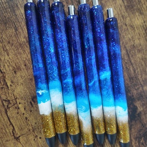 May include: Set of seven blue and gold glitter pens with a white wave design. The pens have silver caps and black tips.