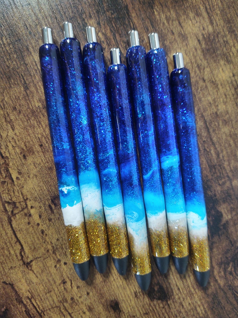 May include: Set of seven blue and gold glitter pens with a white wave design. The pens have silver caps and black tips.