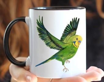 Op de afbeelding: Witte keramische mok met een zwarte rand en handvat. De mok heeft een kleurrijke illustratie van een groene en gele parkiet in vlucht.