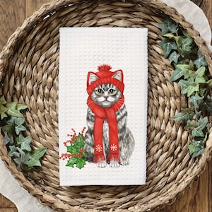 May include: A white kitchen towel with a watercolor illustration of a gray cat wearing a red knit hat and scarf with a red berry sprig. The cat is looking directly at the viewer.