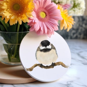 Könnte beinhalten: Ein weißer Untersetzer aus Keramik mit einer Aquarell-Illustration eines Schwarzkappen-Meisen-Vogels, der auf einem Ast sitzt. Der Vogel hat einen grauen Kopf und schwarze Augen, und sein Körper ist weiß mit einem grauen Rücken. Der Untersetzer ist von einem Strauß aus rosa und gelben Gänseblümchen umgeben.