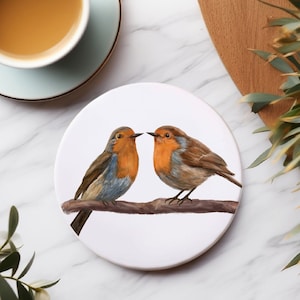 Könnte beinhalten: Ein weißer Untersetzer aus Keramik mit einem gemalten Bild von zwei Rotkehlchen, die auf einem Ast sitzen. Die Rotkehlchen schauen sich an und haben orangefarbene Brüste, braune Flügel und graue Köpfe.