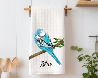 Op de afbeelding: Een witte keukendoek met een blauwe en witte parkiet die op een tak zit met oranje bloesems. De tekst "Blue" is onder de vogel gedrukt.