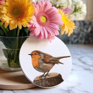 May include: A white ceramic coaster featuring a hand-painted image of a robin perched on a tree stump. The robin has a bright orange breast and a brown back. The coaster is surrounded by a bouquet of pink and yellow flowers.