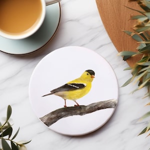 Könnte beinhalten: Ein runder Untersetzer aus Keramik mit weißem Hintergrund, auf dem ein gelber und schwarzer Vogel auf einem Ast sitzt. Der Vogel hat einen schwarzen Kopf und einen gelben Körper mit schwarzen Streifen.