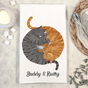 May include: White kitchen towel with a gray and orange cat illustration. The cats are curled up together in a yin yang shape. The text "Buddy & Rusty" is printed below the cats.