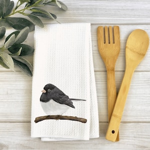May include: White kitchen towel with a black and white bird illustration on a branch. The bird is perched on a branch with brown leaves. The towel is folded and placed on a wooden surface with a wooden spoon and fork.
