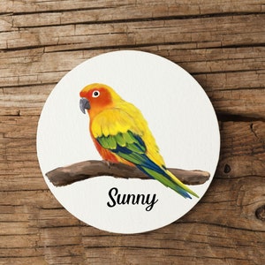 Könnte beinhalten: Ein runder, weißer Untersetzer mit einer Aquarellillustration eines lebhaften gelb, orange und grün gefärbten Papageis, der auf einem braunen Ast sitzt. Das Wort "Sunny" steht in schwarzer Schrift darunter. Der Untersetzer befindet sich vor einem Holzgrund.