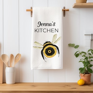 Può includere: Asciugamano da cucina bianco con un'illustrazione di un'ape nera e gialla e il testo "Jenna's Kitchen" stampato in nero.