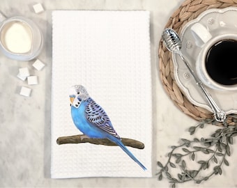 Op de afbeelding: Een witte keukendoek met een blauwe en witte parkiet die op een tak zit. De parkiet heeft een gele snavel en een wit hoofd.