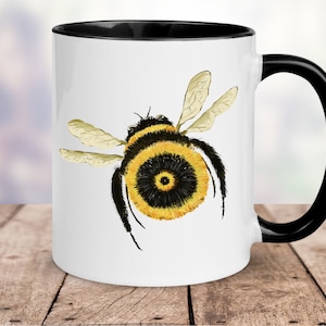 Può includere: Tazza in ceramica bianca con bordo e manico neri. La tazza presenta un'illustrazione ad acquerello di un bombo con strisce gialle e nere.