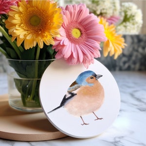 May include: A white ceramic coaster featuring a hand-painted illustration of a blue and pink bird. The bird is perched on a branch and has a long tail. The coaster is surrounded by a bouquet of pink, yellow and white flowers.