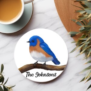 Könnte beinhalten: Ein weißer Untersetzer aus Keramik mit einem blauen Vogel, der auf einem Ast sitzt. Der Vogel hat blaue Flügel, eine weiße Brust und einen orangefarbenen Bauch. Der Text "The Johnsons" ist unter dem Vogel gedruckt.