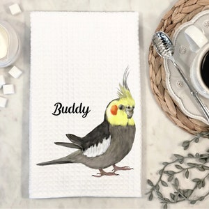 Puede incluir: Toalla de cocina blanca con una ilustración de un cacatúa amarillo y gris. El pájaro está mirando hacia la derecha y tiene una cabeza amarilla y un cuerpo gris. El texto "Buddy" está impreso en negro debajo del pájaro.