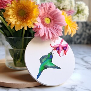 May include: A white round coaster featuring a green hummingbird with a long beak drinking nectar from a pink flower. The coaster is resting on a light brown surface.