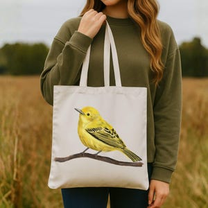 May include: A cream-colored tote bag featuring a detailed illustration of a yellow bird perched on a brown branch. The bird has black markings on its wings and tail. The bag has white straps and is held by a person wearing a green sweatshirt.