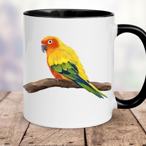 May include: A white ceramic mug with a black rim and handle features a colourful illustration of a yellow, orange, green, and blue parrot perched on a branch.