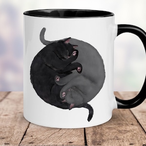 May include: White ceramic mug with a black rim and handle. The mug features a black and gray cat design in the shape of a yin yang symbol.