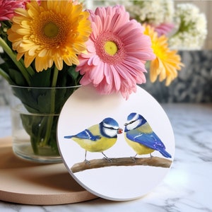 May include: A white ceramic coaster featuring a painted design of two bluebirds perched on a branch. The birds are facing each other, and one is holding a red berry in its beak.