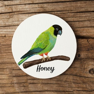 Könnte beinhalten: Ein runder weißer Untersetzer mit einem grün-schwarzen Vogel, der auf einem Ast sitzt. Der Vogel hat einen schwarzen Kopf und einen roten Schnabel. Der Text "Honey" ist unter dem Vogel gedruckt.