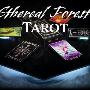 May include: A black box with the text "Ethereal Forest Tarot" on top. The box is open and several cards are spread out on a black surface. The cards feature illustrations of a hummingbird, a snowflake, and a forest scene.
