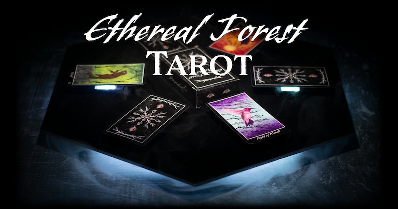May include: A black box with the text "Ethereal Forest Tarot" on top. The box is open and several cards are spread out on a black surface. The cards feature illustrations of a hummingbird, a snowflake, and a forest scene.