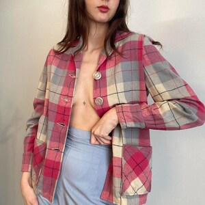 May include: A vintage plaid jacket in shades of pink, gray, and beige. The jacket features a classic button-up design with two front pockets and a collared neckline. The fabric has a woven texture, and the jacket is worn open, revealing a light blue skirt.