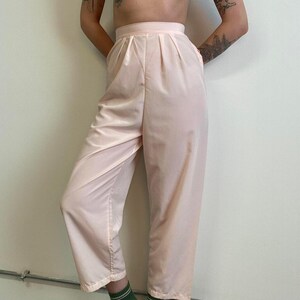 May include: Pale pink high-waisted pants with pleats at the waist and pockets. The pants are made of a smooth, lightweight fabric. The pants are a solid color and have a relaxed fit.